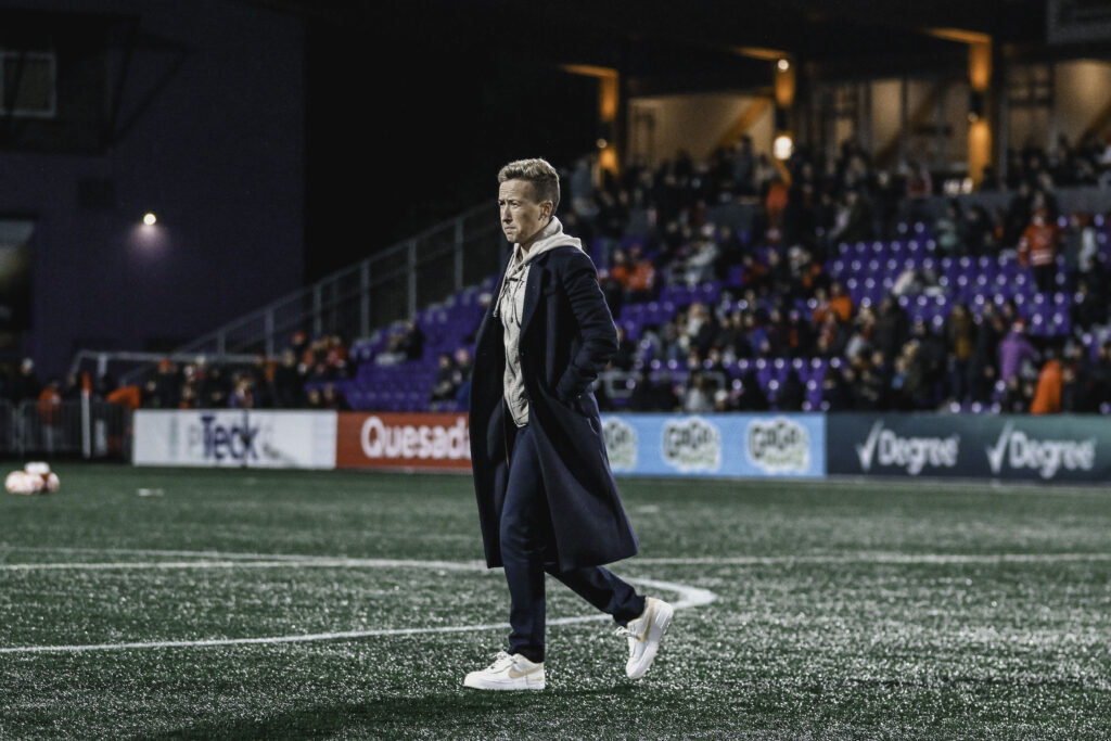 Bev Priestman is seen walking on a soccer field wearing a trenchcoat.